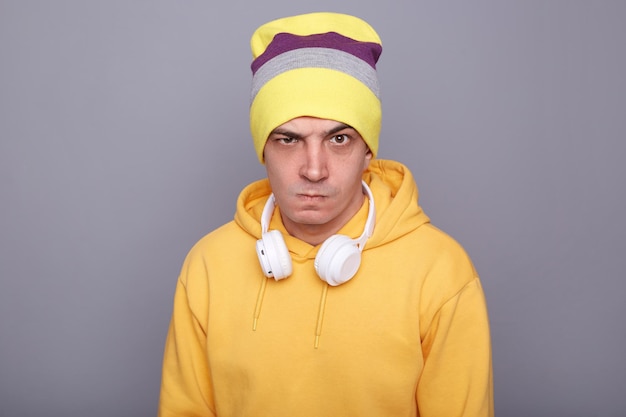 Afbeelding van een boze agressieve blanke hipster man met muts en gele casual hoodie die ruzie maakt met iemand die naar de camera kijkt met opeengeklemde tanden staand geïsoleerd over grijze achtergrond