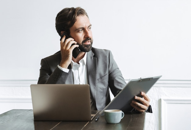 Afbeelding van een denkende zakenman die een formeel pak draagt dat klembord vasthoudt en op zijn mobiel praat terwijl hij op kantoor werkt