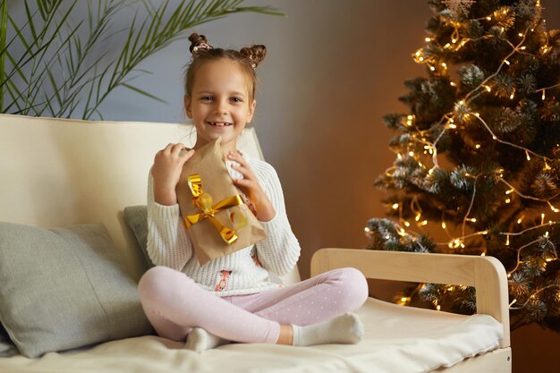 Afbeelding van een extreem gelukkig klein meisje met grappige haarbroodjes met een ingepakte huidige doos kijkend naar de camera met een brede glimlach zittend op de bank in de buurt van de kerstboom thuis