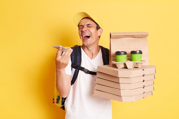 Afbeelding van een extreem gelukkige positieve bezorger met een pakketje met pizza en koffie geïsoleerd op een gele achtergrond met een mobiele telefoon in handen die spraakberichten opneemt van het lachen