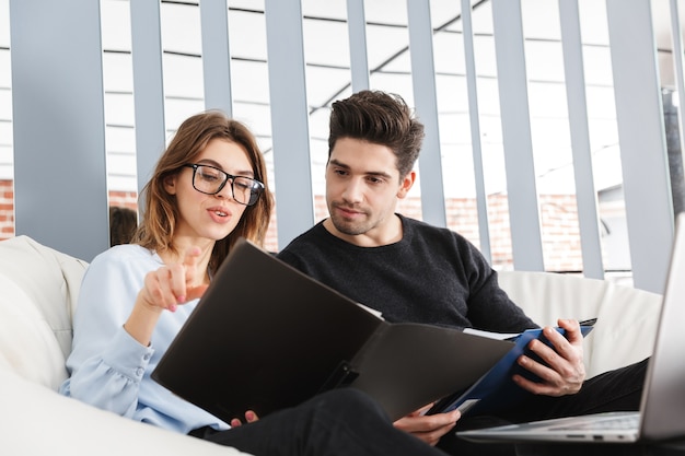 Afbeelding van een geconcentreerde jonge verliefde paar thuis binnenshuis met behulp van laptop computer werken met documenten.