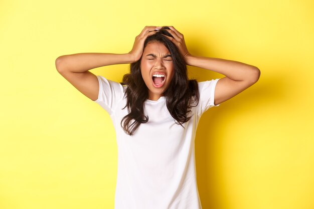 Afbeelding van een gefrustreerde Afro-Amerikaanse vrouw, die er pissig uitziet, schreeuwt en handen op het hoofd houdt, staande over een gele achtergrond.