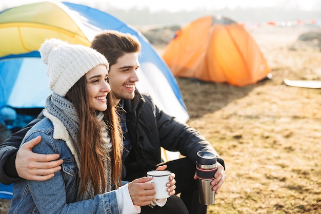 Afbeelding van een gelukkig jong liefdevol paar buiten met een tent in een gratis alternatieve vakantiecamping boven de bergen die hete thee drinken.