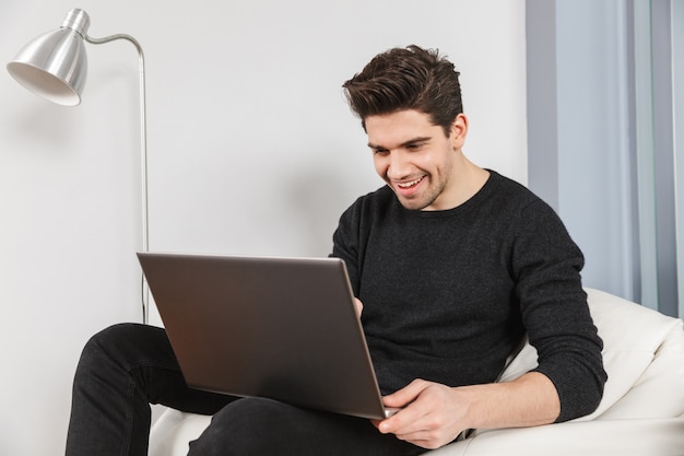 Afbeelding van een gelukkig knappe jonge man thuis binnenshuis met behulp van laptopcomputer.