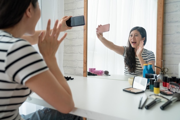 Afbeelding van een gelukkige jonge aziatische vrouw die thuis aan de kaptafel zit en make-up doet met cosmetica. vrolijk meisje met video-telefoongesprek maak je klaar in de ochtend vrolijk zwaaiende hand mobiel