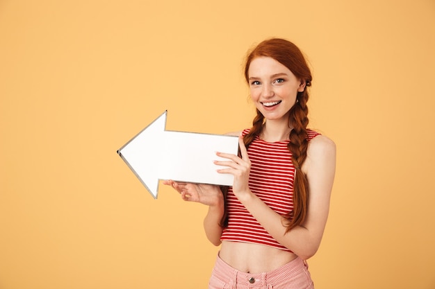 Afbeelding van een gelukkige jonge mooie roodharige vrouw poseren geïsoleerd over gele muur met pijl.