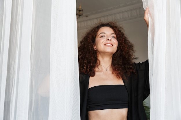 Afbeelding van een gelukkige jonge vrolijke vrouw in lingerie badmode poseren binnenshuis in huishotel in de buurt van gordijn.