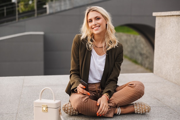 Afbeelding van een gelukkige jonge vrouw buiten zitten kijken met behulp van telefoon.