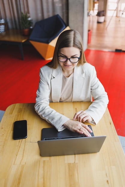 Afbeelding van een gelukkige jonge vrouw in een jas die lacht en op een laptop werkt terwijl ze telefoneert in een modern kantoor met grote ramen Werk op afstand