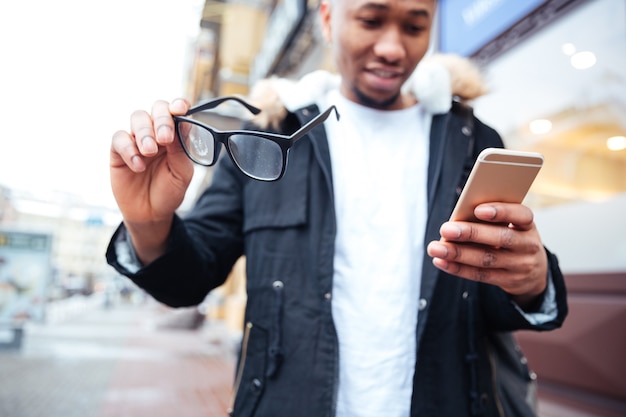 Afbeelding van een gelukkige man die zijn bril en telefoon in handen houdt terwijl hij buiten loopt. Kijk naar telefoon.