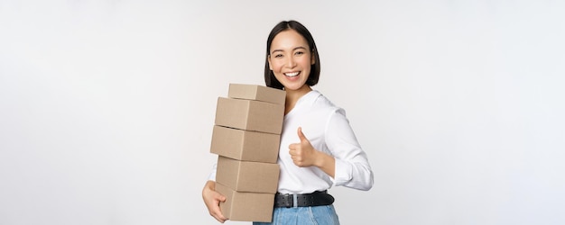 Afbeelding van een gelukkige moderne aziatische vrouw die duimen laat zien terwijl ze dozen leveringsgoederen vasthoudt die tegen een witte achtergrond staan