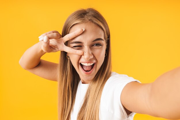 Afbeelding van een gelukkige vrouw in eenvoudige kleding die lacht en een vredesteken toont terwijl ze een selfie-foto maakt die over een gele muur is geïsoleerd