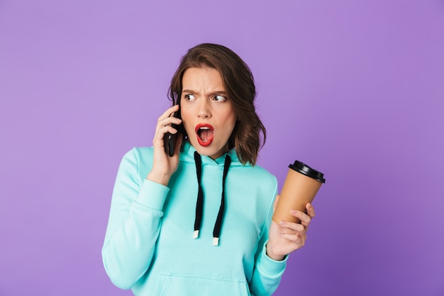 Afbeelding van een geschokt ontevreden jonge vrouw poseren geïsoleerd over paarse muur muur praten door mobiele telefoon.