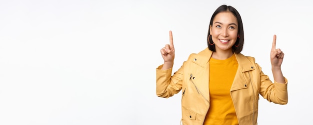 Afbeelding van een glimlachende Aziatische brunette vrouw die met haar vingers omhoog wijst en reclame toont met een blij gezicht dat zich voordeed tegen een witte achtergrond