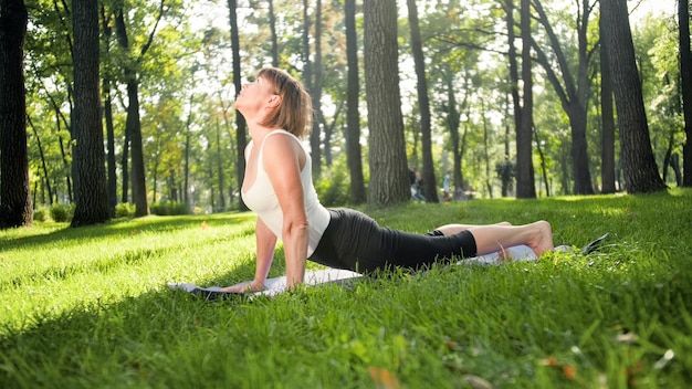 Afbeelding van een glimlachende gelukkige vrouw van middelbare leeftijd die mediteert en yoga-oefeningen doet op gras in het bos. Vrouw die voor haar fysieke en mentale gezondheid zorgt terwijl ze aan fitness doet en zich uitstrekt in het park