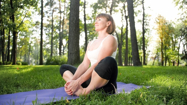 Afbeelding van een glimlachende gelukkige vrouw van middelbare leeftijd die mediteert en yoga-oefeningen doet op gras in het bos. Vrouw die voor haar fysieke en mentale gezondheid zorgt terwijl ze aan fitness doet en zich uitstrekt in het park