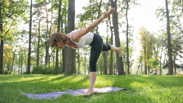 Afbeelding van een glimlachende gelukkige vrouw van middelbare leeftijd die mediteert en yoga-oefeningen doet op gras in het bos. Vrouw die voor haar fysieke en mentale gezondheid zorgt terwijl ze aan fitness doet en zich uitstrekt in het park