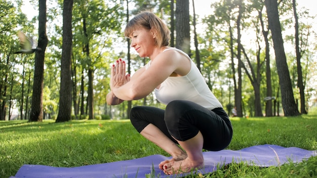Afbeelding van een glimlachende vrouw van middelbare leeftijd in fitnesskleding die rek- en yogaoefeningen doet. Vrouw mediteren en sporten op fitnessmat op gras in het park