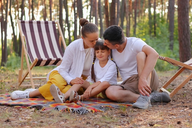 Afbeelding van een jong gezin dat geniet van een moment terwijl ze op een deken in het park zitten en hun schattige dochter kussen, mensen die ontspannen in het hout en samen een weekend doorbrengen