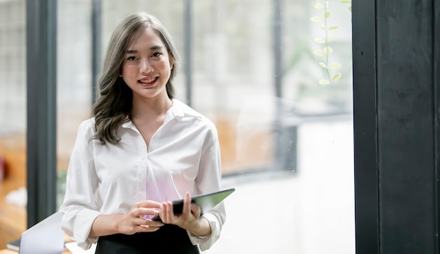 Afbeelding van een jonge aziatische vrouw die in het bedrijf lacht en een digitale tablet vasthoudt die in een helder kantoor staat