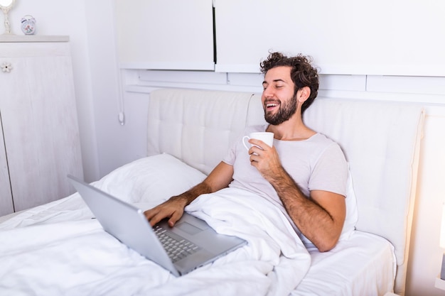 Afbeelding van een jonge blanke man drinkt koffie in de ochtend, zit op bed, werkt op zijn laptop en drinkt koffie, bereidt zich voor op onderzoek, gebruikt een laptop om op internet te surfen.