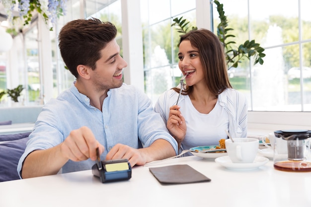 Afbeelding van een jonge gelukkige verliefde paar zitten in de creditcard van de caféholding en betalen.