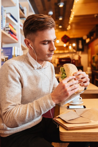 Afbeelding van een jonge knappe man gekleed in een wit overhemd die in een café zit terwijl hij naar muziek luistert en een broodje vasthoudt.