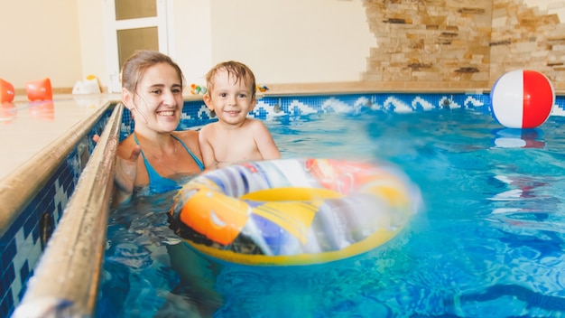Afbeelding van een jonge moeder die haar kleine 3-jarige jongenskind leert zwemmen en met een kleurrijke strandbal speelt in het binnenzwembad indoor