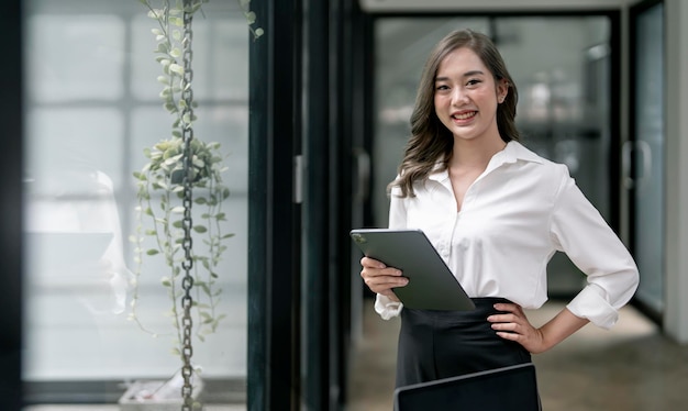 Afbeelding van een jonge smiley-aziatische vrouw die in het bedrijf lacht en een digitale tablet vasthoudt die in een helder kantoor staat