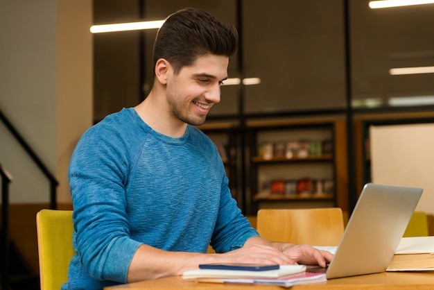Afbeelding van een jonge student gelukkig man in bibliotheek huiswerk studeren lezen en met behulp van laptopcomputer.