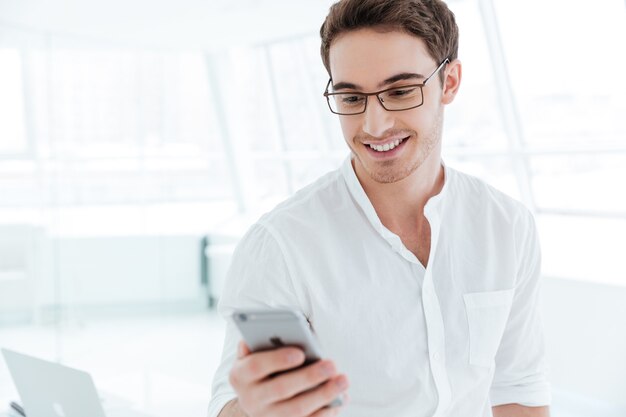 Afbeelding van een jonge vrolijke man gekleed in een wit overhemd die telefonisch aan het chatten is. Telefoon kijken.