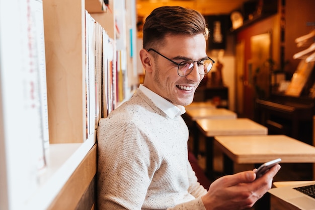 Afbeelding van een jonge vrolijke man met een bril die in café zit terwijl hij een mobiele telefoon gebruikt.