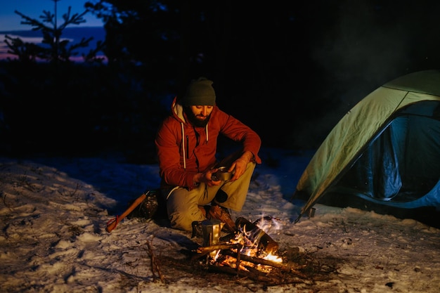 Afbeelding van een jonge wandelaar die een diner kookt bij een kampvuur in de bergen in de winter Mannelijke wandelaar met een baard die na zonsondergang bij de tent zit en een kampvuur maakt na het wandelen