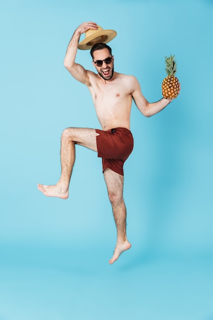 Afbeelding van een knappe jonge zomergelukkige man die geïsoleerd over een blauwe muur springt met ananas.