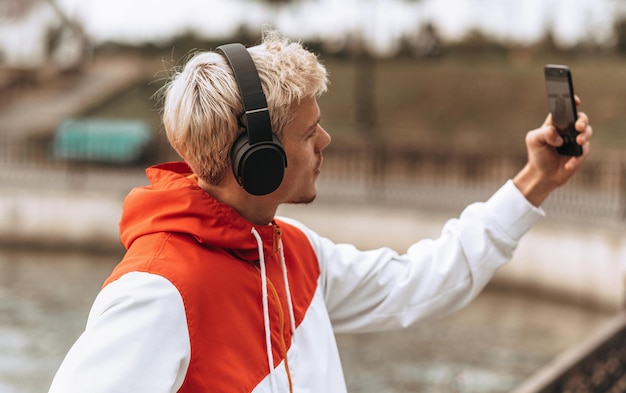 Foto afbeelding van een knappe jongeman die een zelfportret maakt op haar smartphone in het stadspark mannelijke student die slefie maakt terwijl hij naar zijn favoriete muziek luistert via een draadloze hoofdtelefoon buiten