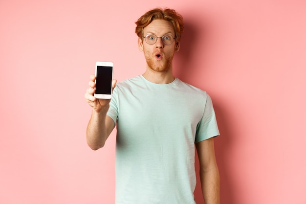Afbeelding van een knappe man met rood haar, een bril en een t-shirt, die wauw zegt en het smartphonescherm laat zien, staande tegen een roze achtergrond.