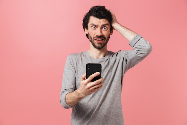 Afbeelding van een knappe verwarde ernstige jonge man over roze muur met behulp van mobiele telefoon.