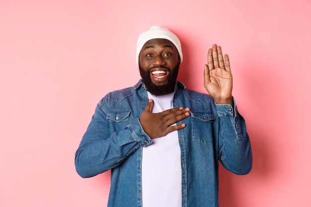 Afbeelding van een lachende Afro-Amerikaanse man die eerlijk is, de waarheid vertelt, handen op het hart en in de lucht, belofte doen of vloeken, staande op een roze achtergrond.