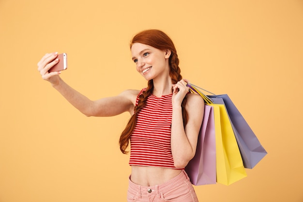 Afbeelding van een lachende jonge mooie roodharige vrouw die zich geïsoleerd over een gele muur met boodschappentassen poseert, neemt een selfie via de mobiele telefoon.