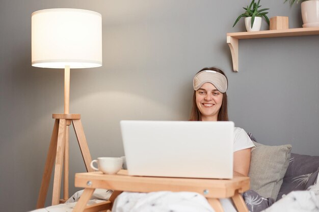 Afbeelding van een lachende mooie bruinharige vrouw in een wit t-shirt en geblinddoekt zittend in bed voor een laptop kijkend naar een film kijkend naar een film die vrolijk glimlacht