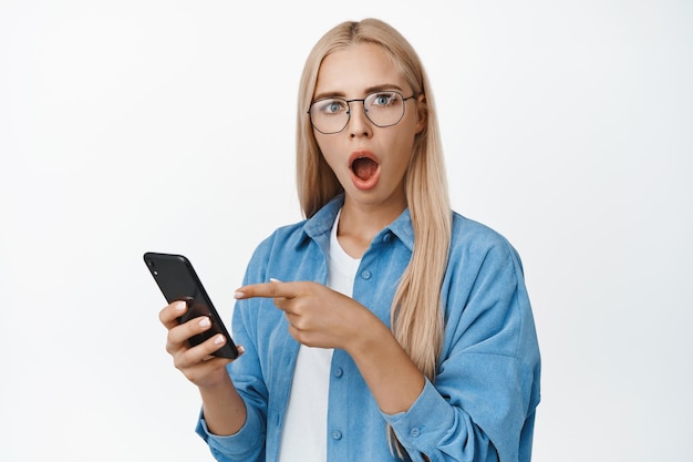 Afbeelding van een meisje met een bril houdt de vinger van de telefoon vast op de smartphone met een verrast en geschokt gezicht tegen een witte achtergrond