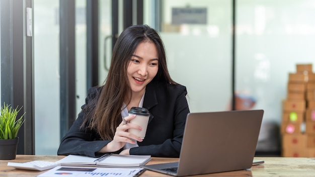 Afbeelding van een mooie Aziatische zakenvrouw gelukkig een succesvol werk met een koffiekopje met een laptop en document op kantoor.