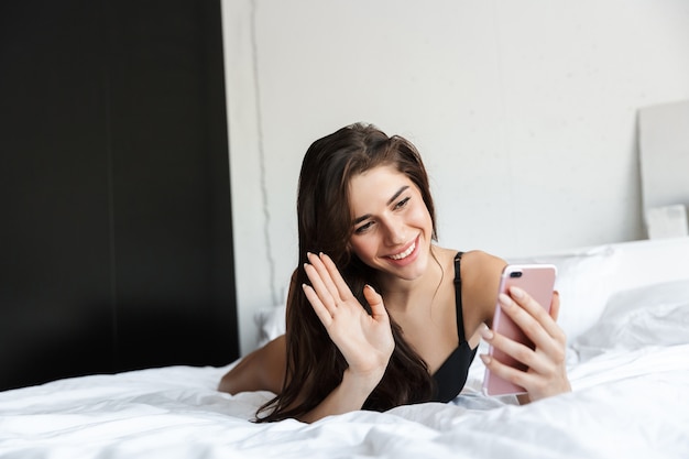 Afbeelding van een mooie brunette vrouw, gekleed in lingerie met behulp van mobiele telefoon praten ligt in bed.