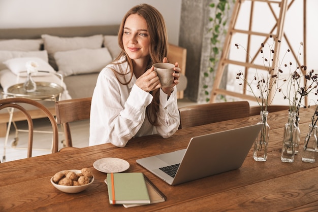 Afbeelding van een mooie jonge glimlachende optimistische gembervrouw die binnenshuis op kantoor zit met een laptopcomputer die koffie drinkt.