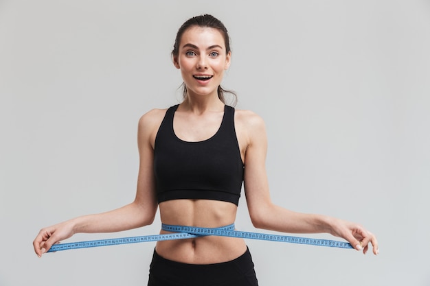 Afbeelding van een mooie jonge sport fitness opgewonden gelukkige vrouw poseren met centimeter over taille geïsoleerd over grijze muur.