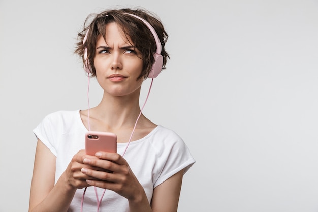 Afbeelding van een mooie jonge, vrij verwarde vrouw die zich voordeed over een witte muurmuur die muziek luistert met een koptelefoon die de telefoon gebruikt.