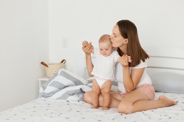 Afbeelding van een mooie moeder, een aantrekkelijke vrouw met een wit overhemd en een korte zitting op bed met haar peuterdochter, mama die haar kind kust en liefde en zachtaardigheid uitdrukt