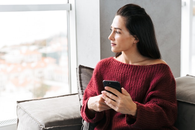 Afbeelding van een mooie vrouw binnenshuis in huis op de bank met behulp van mobiele telefoon.