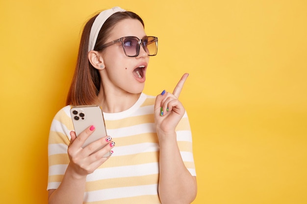 Afbeelding van een opgewonden, verbaasde jonge vrouw met een gestreept t-shirt dat met een mobiele telefoon staat en opzij wijst met de wijsvinger die een gebied toont voor reclame die zich voordeed op een gele achtergrond