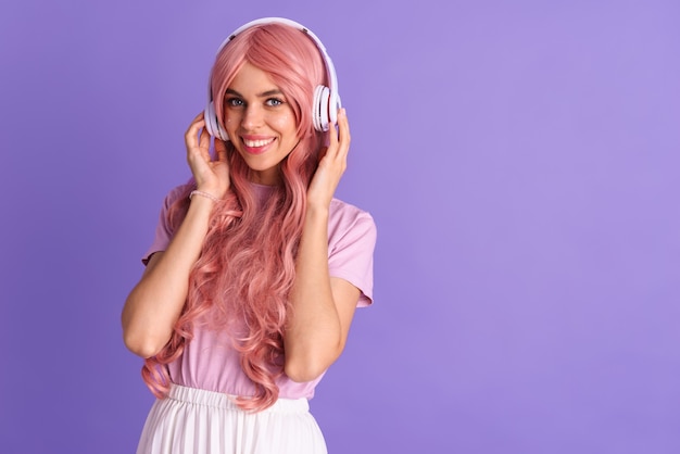 Afbeelding van een optimistisch jong meisje met roze anime-haar poseren geïsoleerd over een paarse muur die muziek luistert met een koptelefoon.